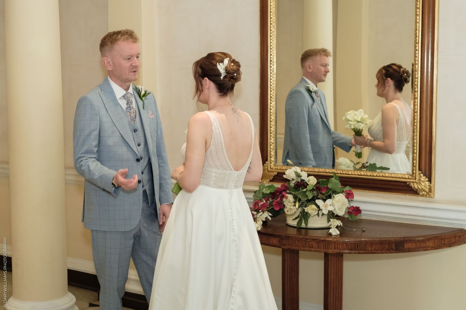 Bride and groom share a tender moment in an elegant hall, with their reflection visible in a ornate gold mirror.