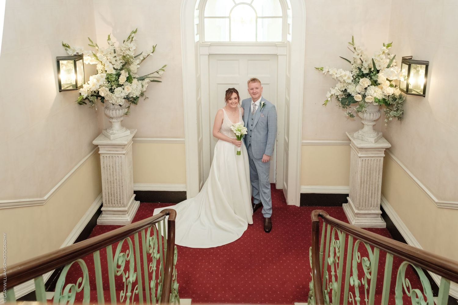 Bride and groom posing on red-carpeted staircase flanked by white floral arrangements in elegant wedding venue.