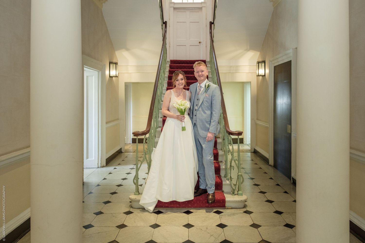 Bride and groom posing together in an elegant hallway with a red-carpeted staircase on their wedding day.