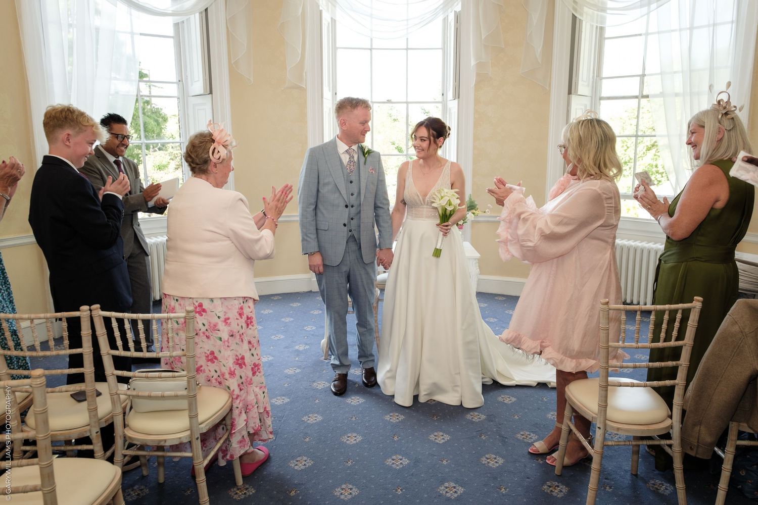 Bride and groom share first dance as newlyweds, surrounded by applauding guests in elegant blue wedding venue.