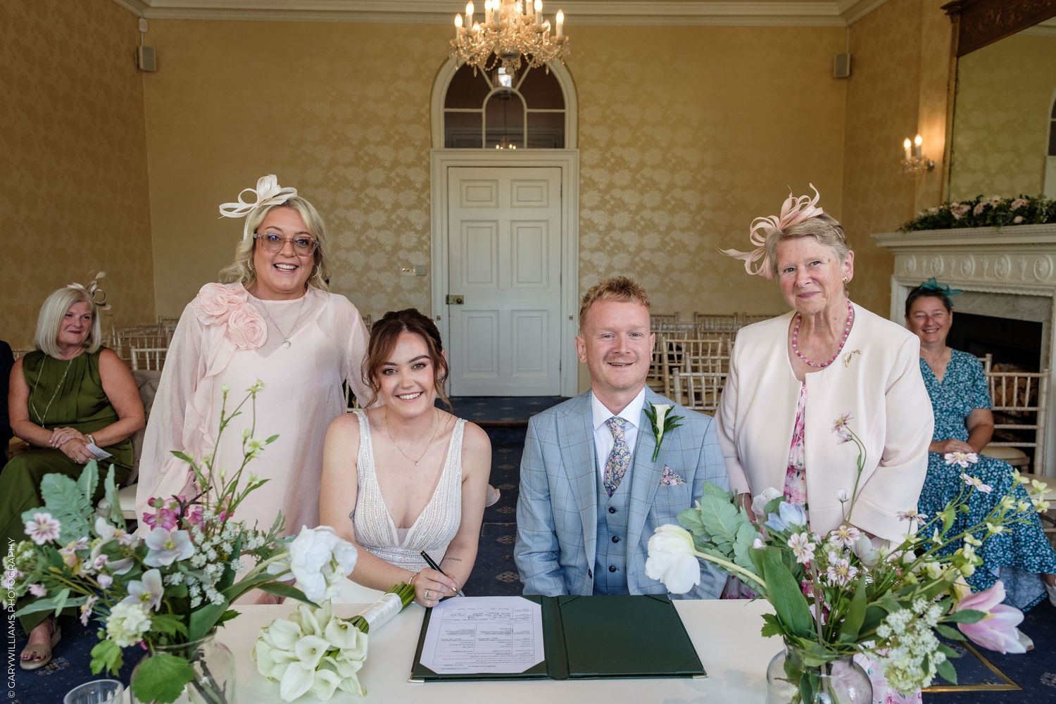 Bride and groom signing wedding register with two female guests in elegant fascinators at a formal venue.