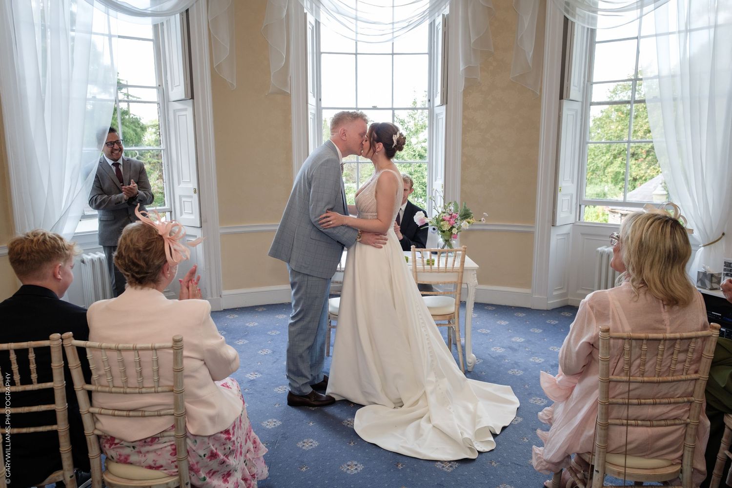 Bride and groom share first kiss at elegant wedding ceremony in a bright room with large windows and seated guests.