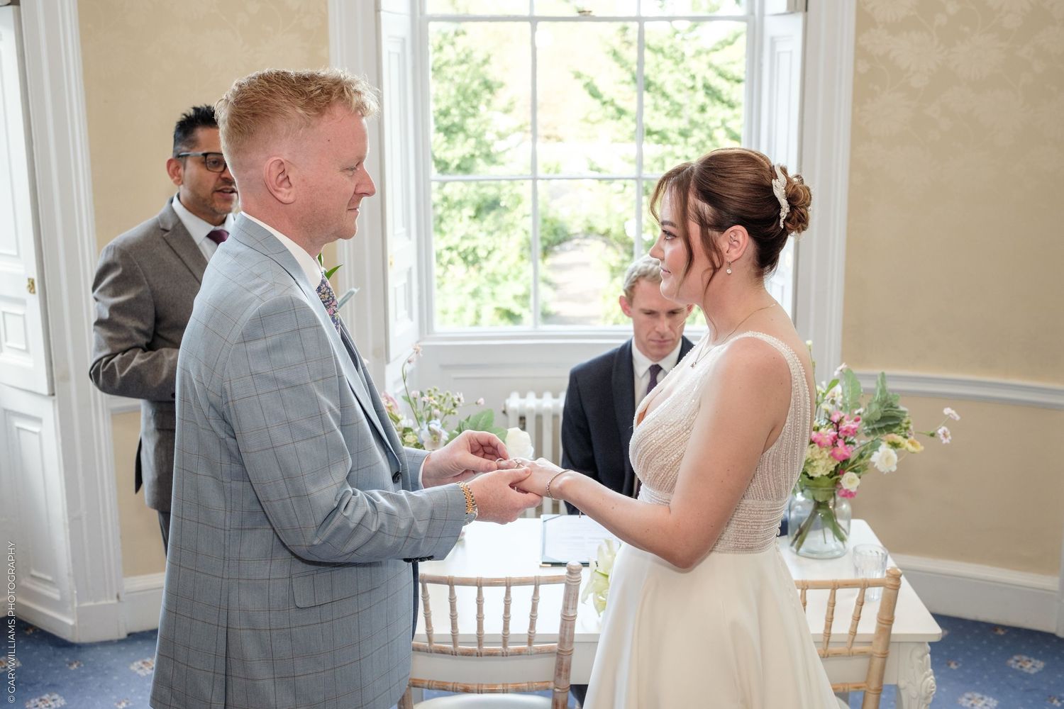 A groom places a wedding ring on his bride's finger during an elegant indoor ceremony with guests watching.
