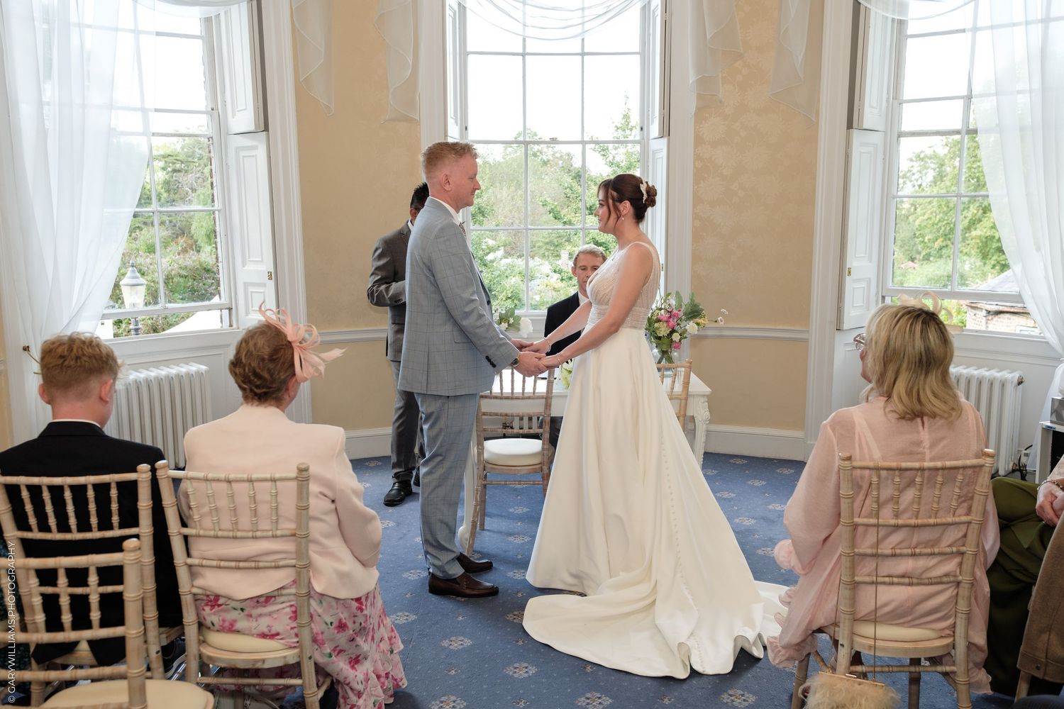 Bride and groom exchange vows at elegant indoor wedding ceremony with guests seated in a blue-carpeted room.