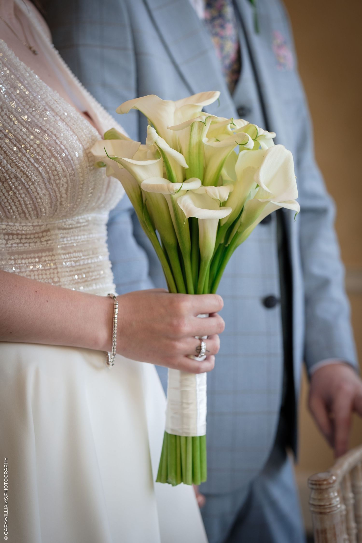 Bride holding elegant white calla lily wedding bouquet with green stems, wearing diamond ring and bracelet.