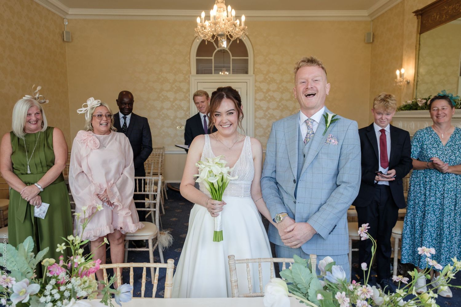 Joyful bride and groom laughing at wedding ceremony with guests and floral arrangements in elegant venue.