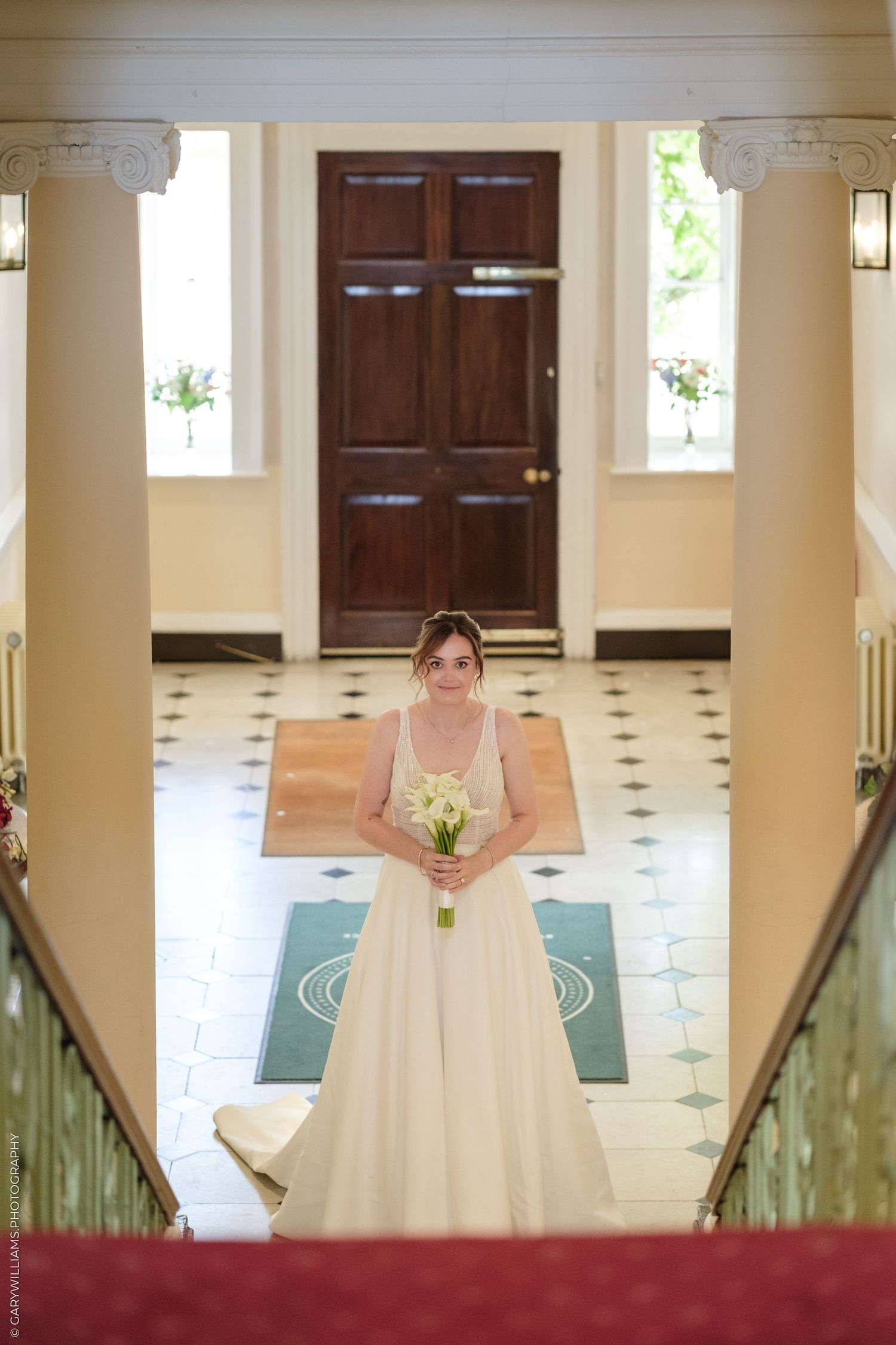 Bride in white gown holding bouquet stands in elegant venue foyer with checkered floor and grand wooden door.