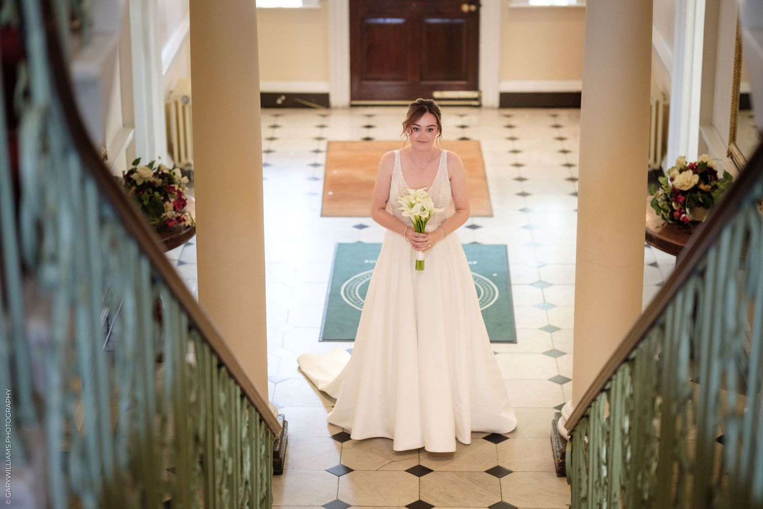 Bride in white wedding gown holding bouquet stands at bottom of elegant staircase with green railings in ornate venue.