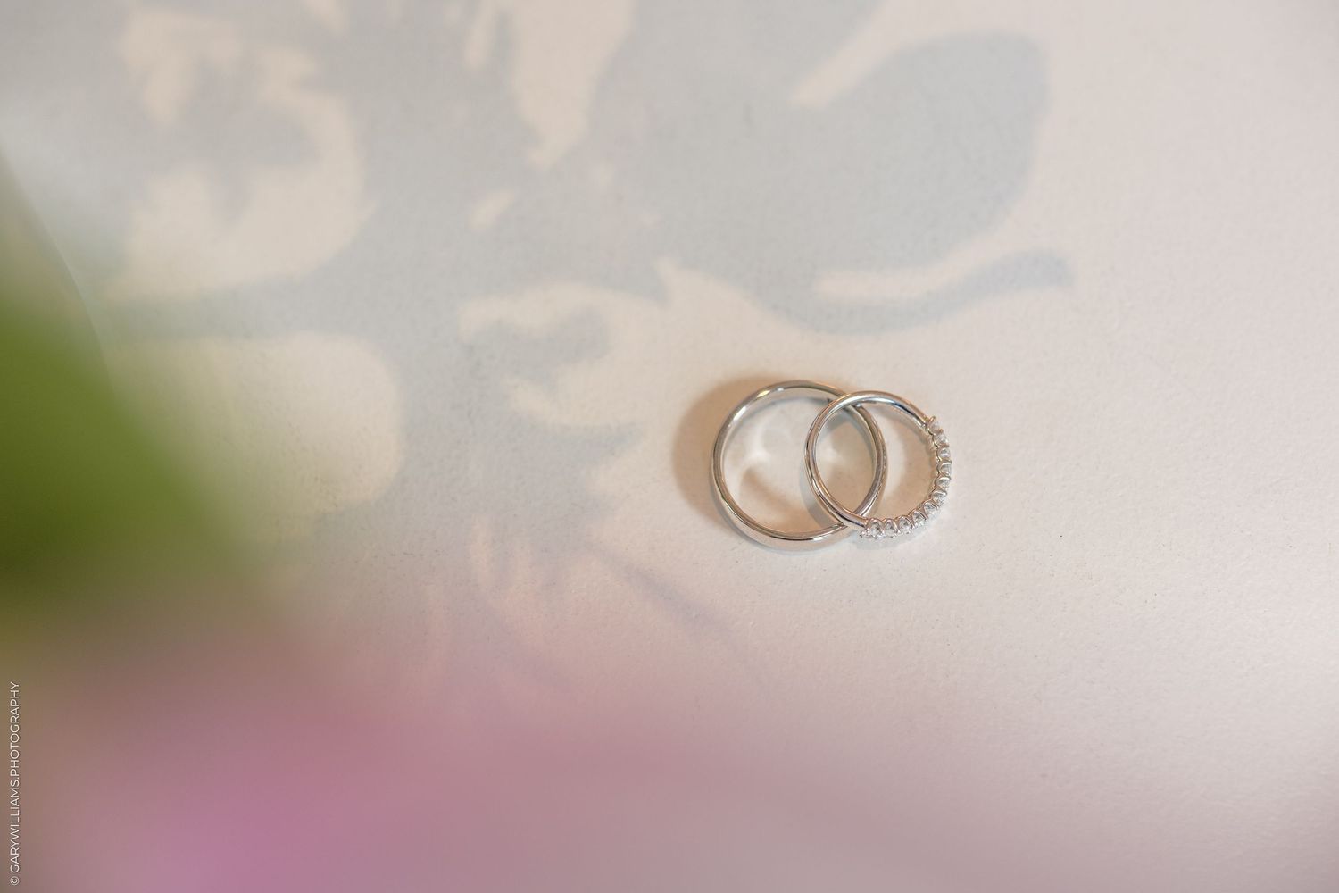 Two silver wedding bands with diamond accents resting together on a soft white surface with floral shadows.