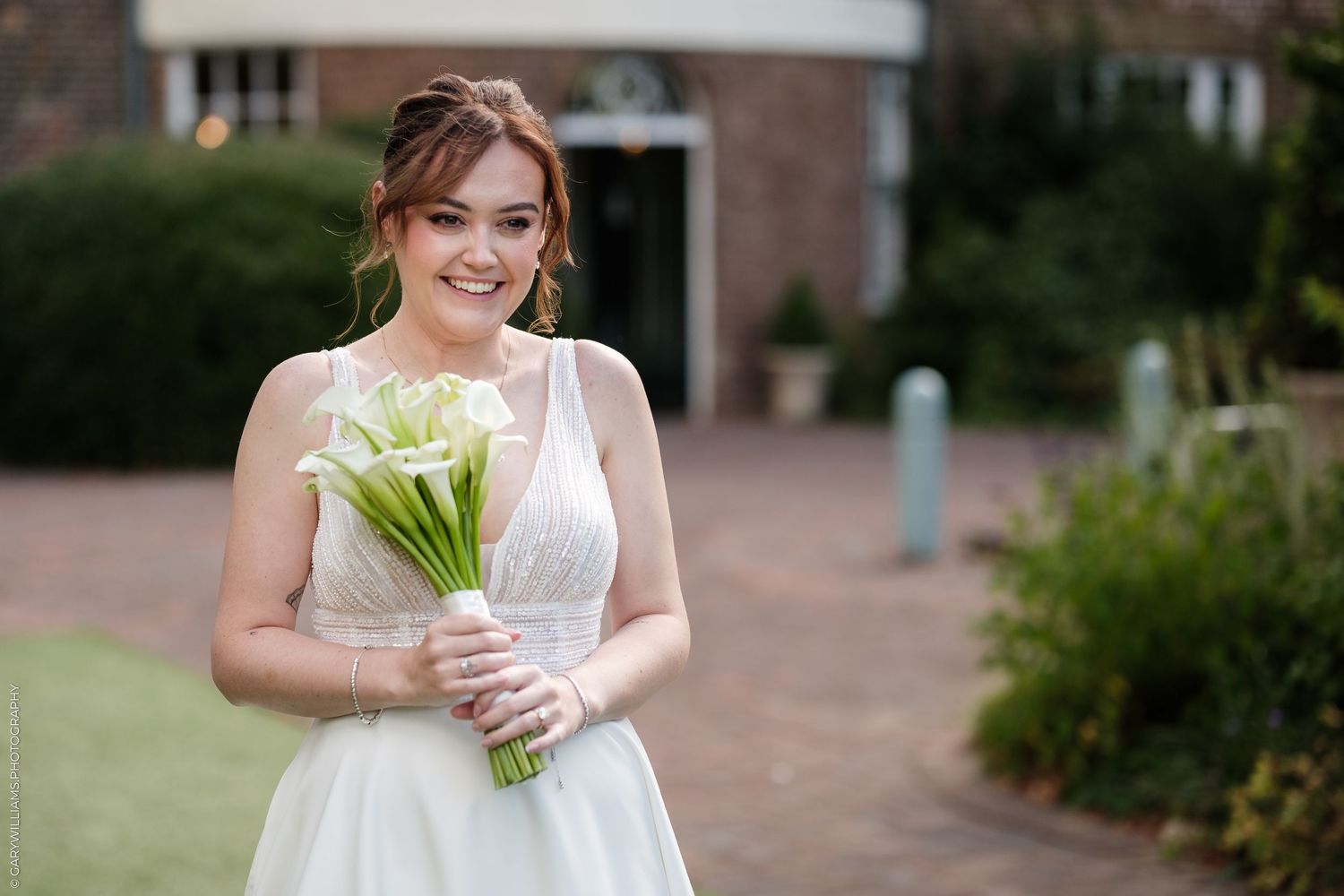 Smiling bride in white gown holding elegant white calla lily bouquet outdoors at wedding venue.