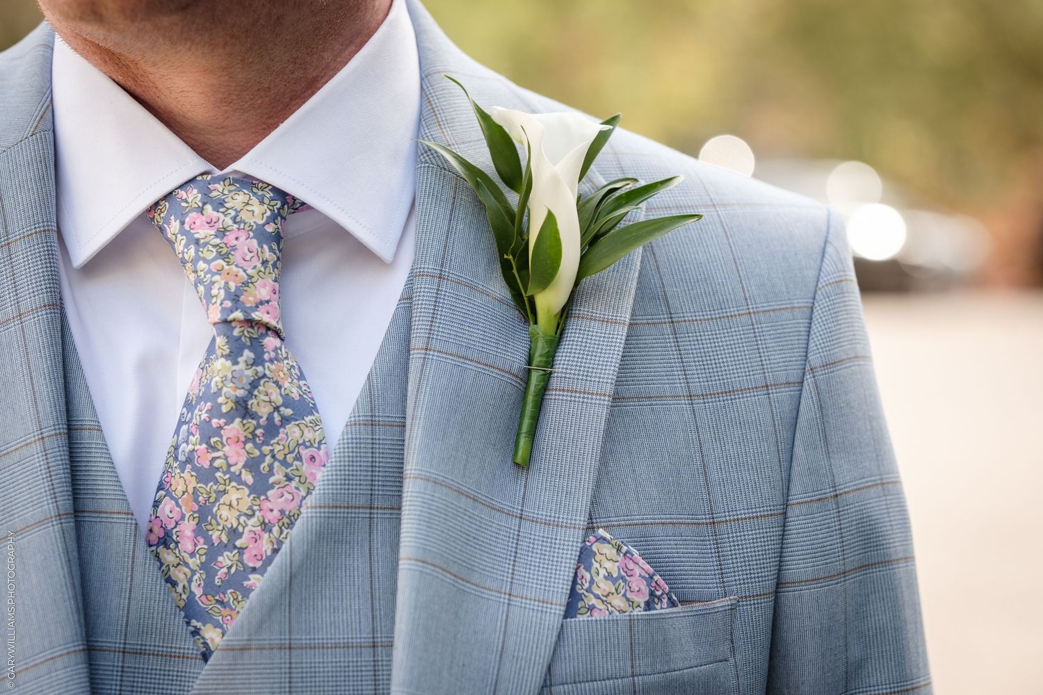 Man in light blue plaid suit with floral tie and white calla lily boutonniere on lapel with matching pocket square.
