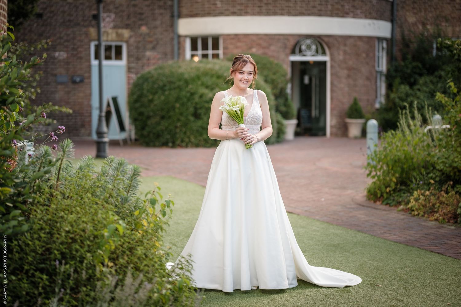 Bride in white gown holding bouquet stands in lush garden outside brick building on her wedding day.