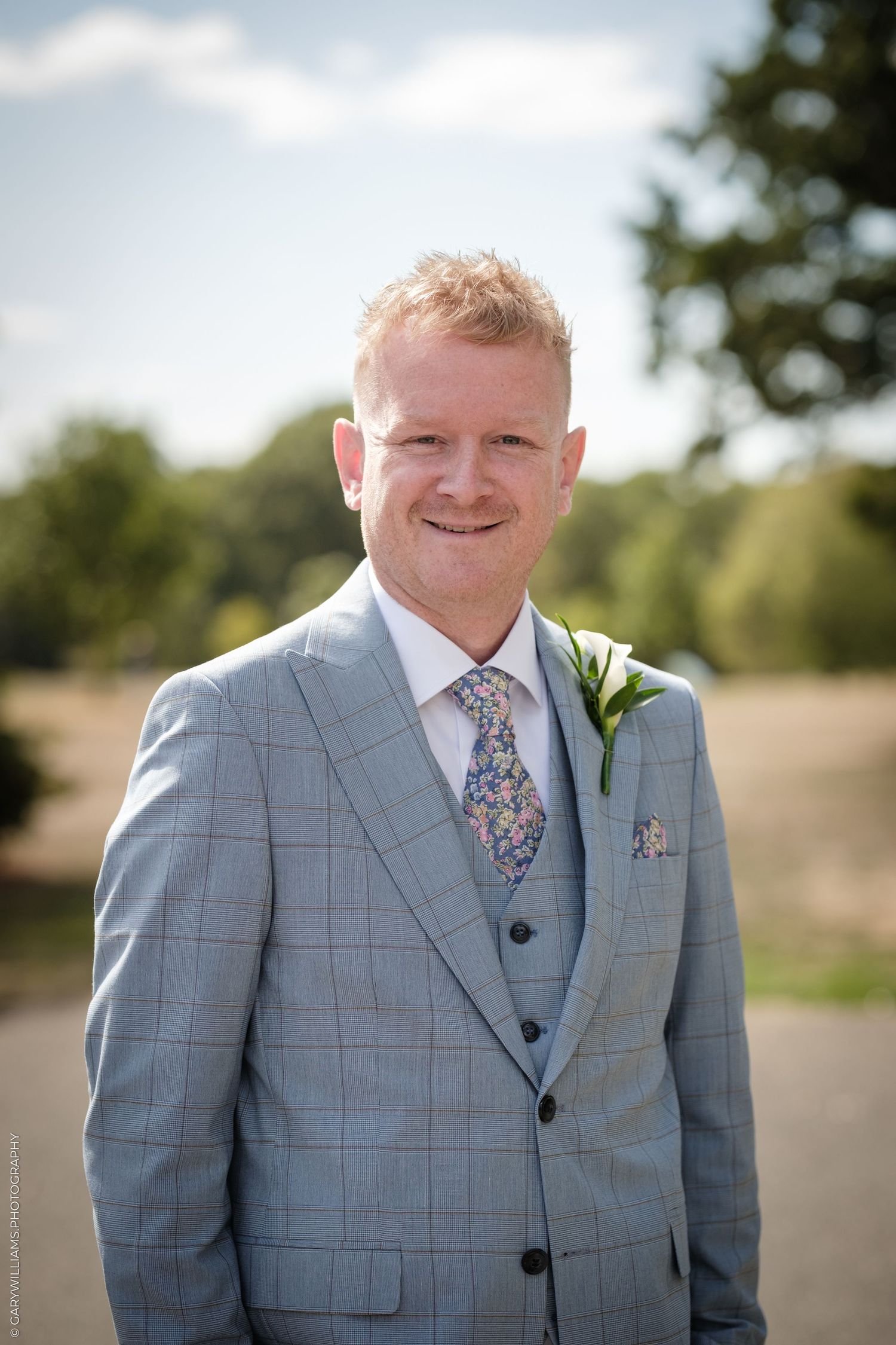 Smiling groom in light blue checked three-piece suit with floral tie and boutonniere outdoors.