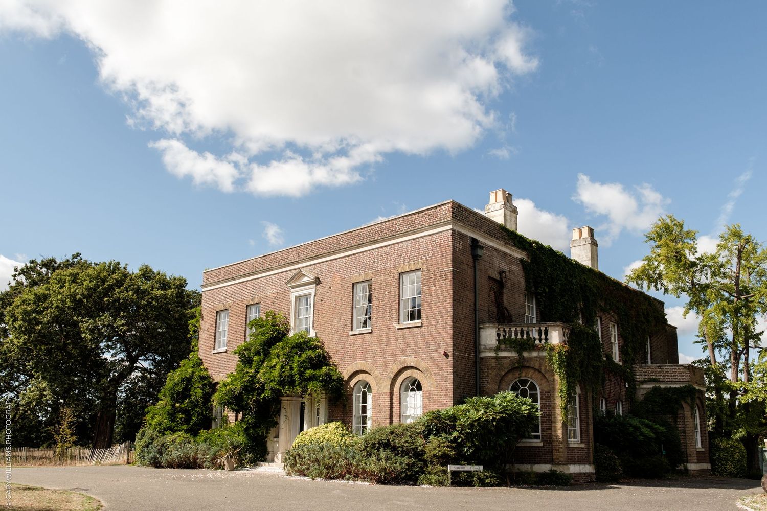 A grand Georgian brick manor house with ivy-covered walls, arched windows, and manicured grounds under a blue sky.