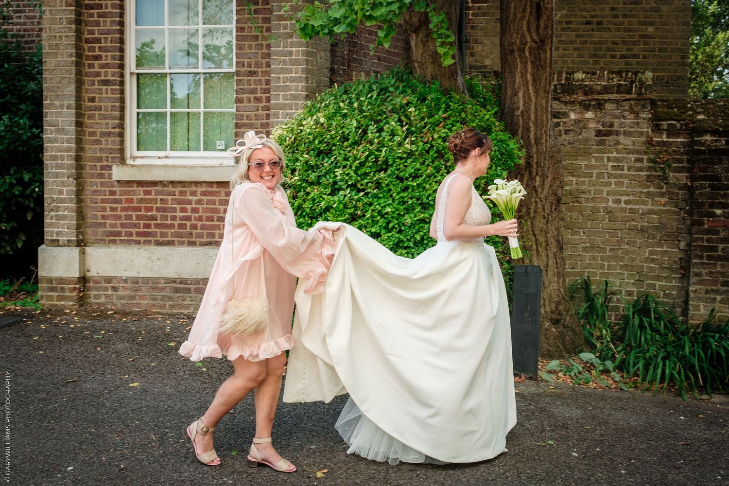 A bridesmaid in pink helps carry the bride's white wedding gown train outside a brick building.