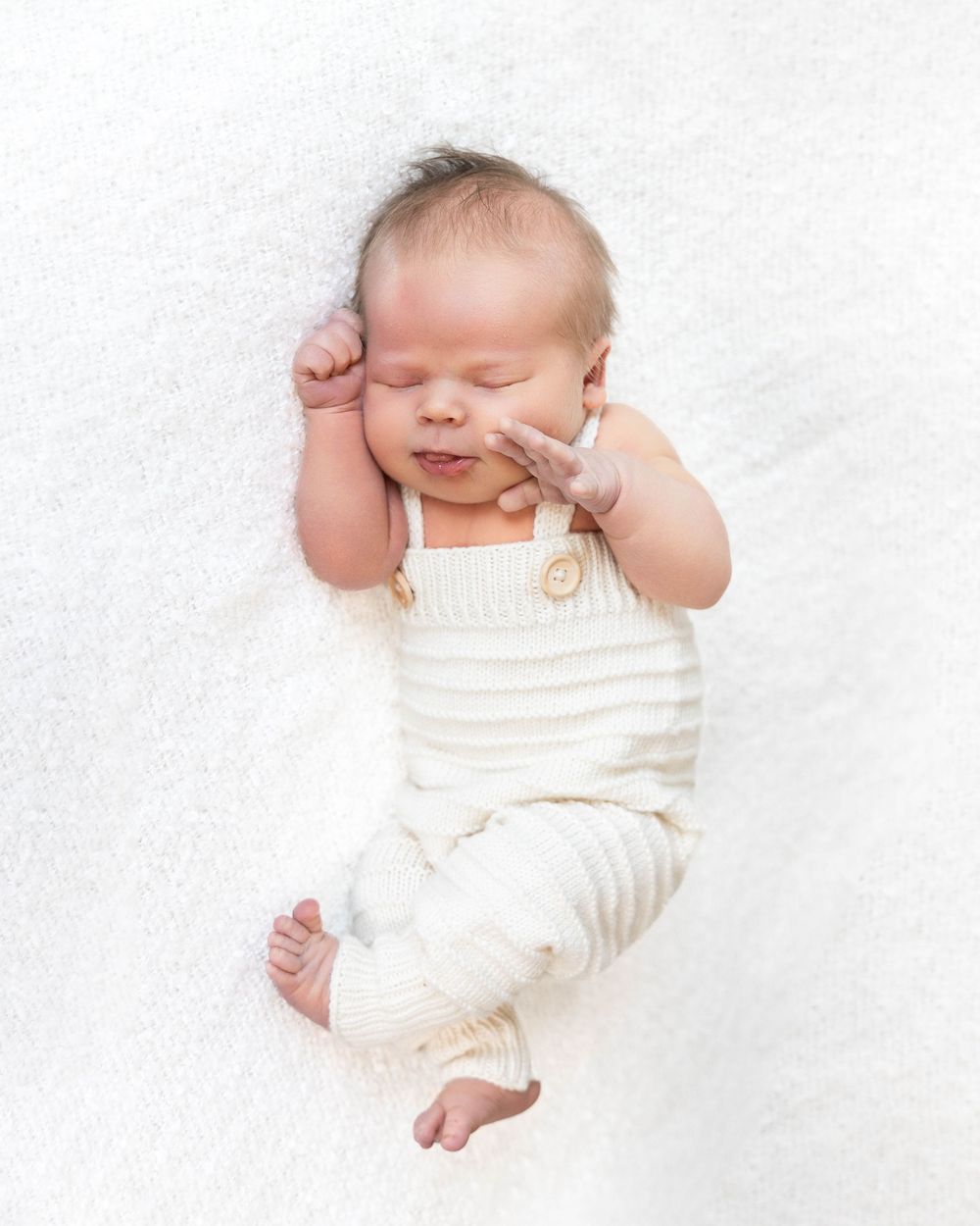 Peaceful newborn baby sleeping in cream knit overalls on a white blanket, with tiny hand raised near face.