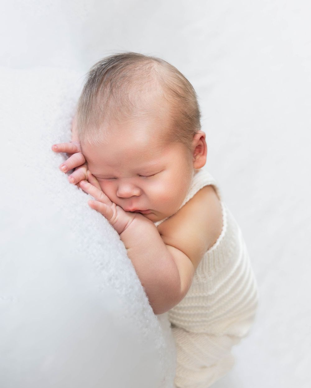 Peaceful newborn baby sleeping on white blanket, hands tucked under cheek, wearing cream knit romper.
