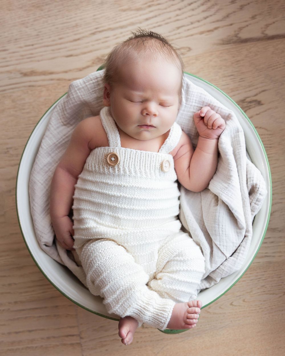 Sleeping newborn baby in cream knit overalls curled in a round bowl with soft white blanket on wood floor.