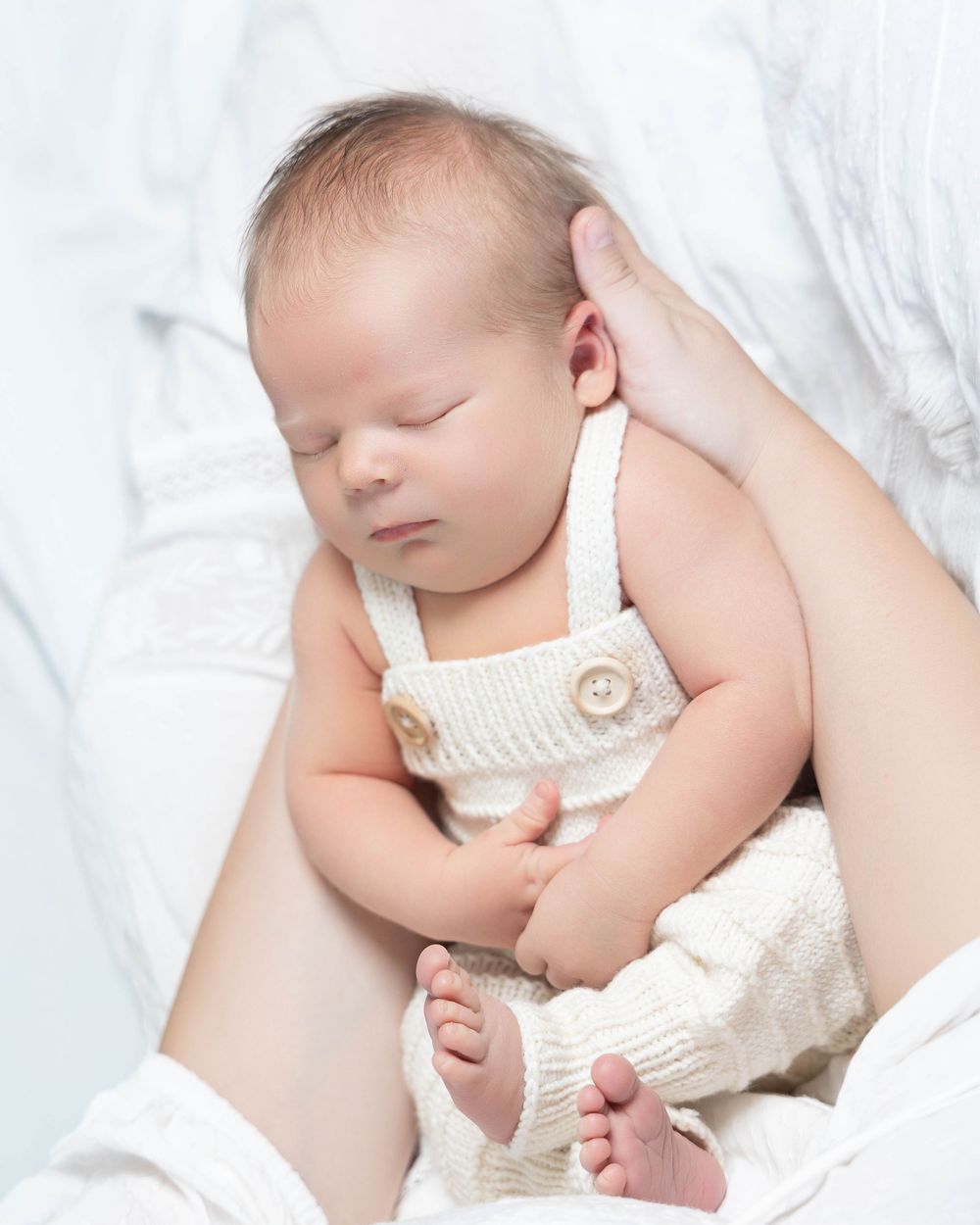 Newborn baby sleeping peacefully in cream knit overalls, cradled in a parent's arms against a soft white background.