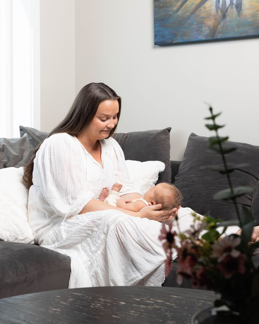 A mother in a white robe sits on a gray sofa, tenderly cradling her newborn baby in a bright, modern living room.