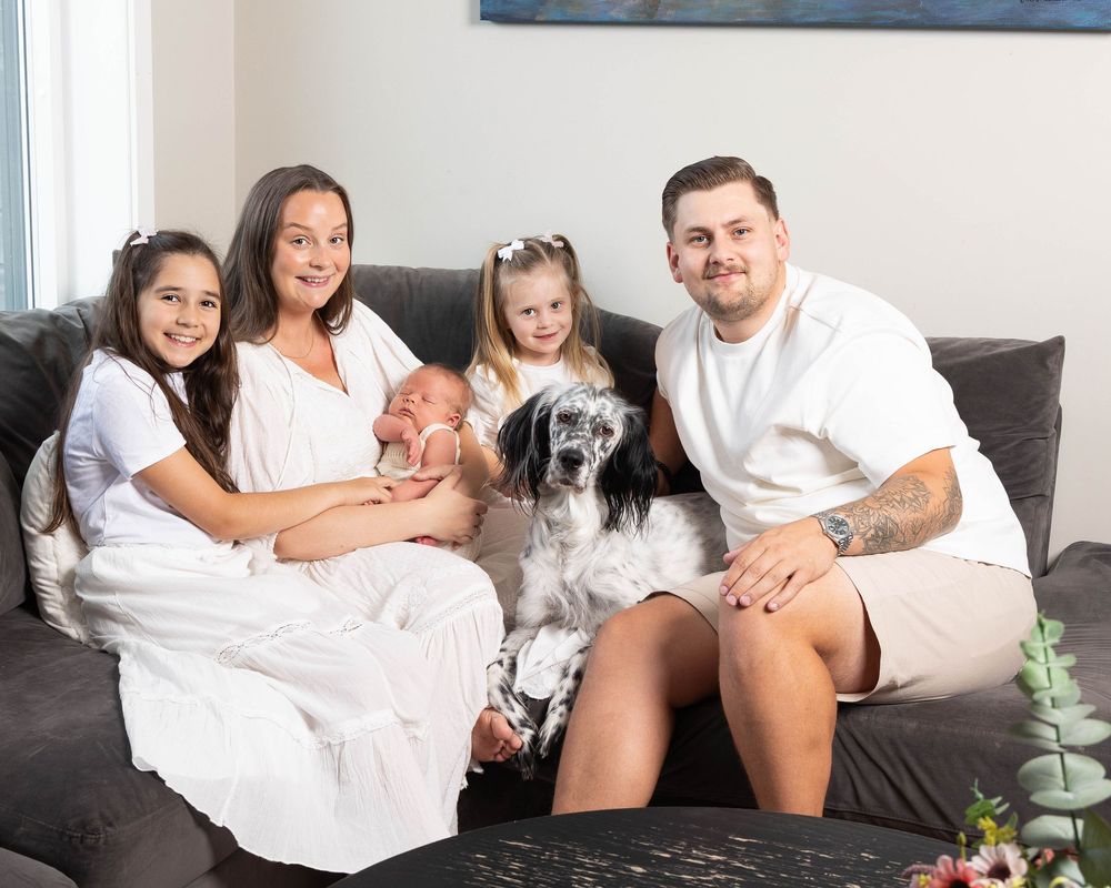 Family of four with newborn baby and spotted dog posing together on gray sofa in bright living room.