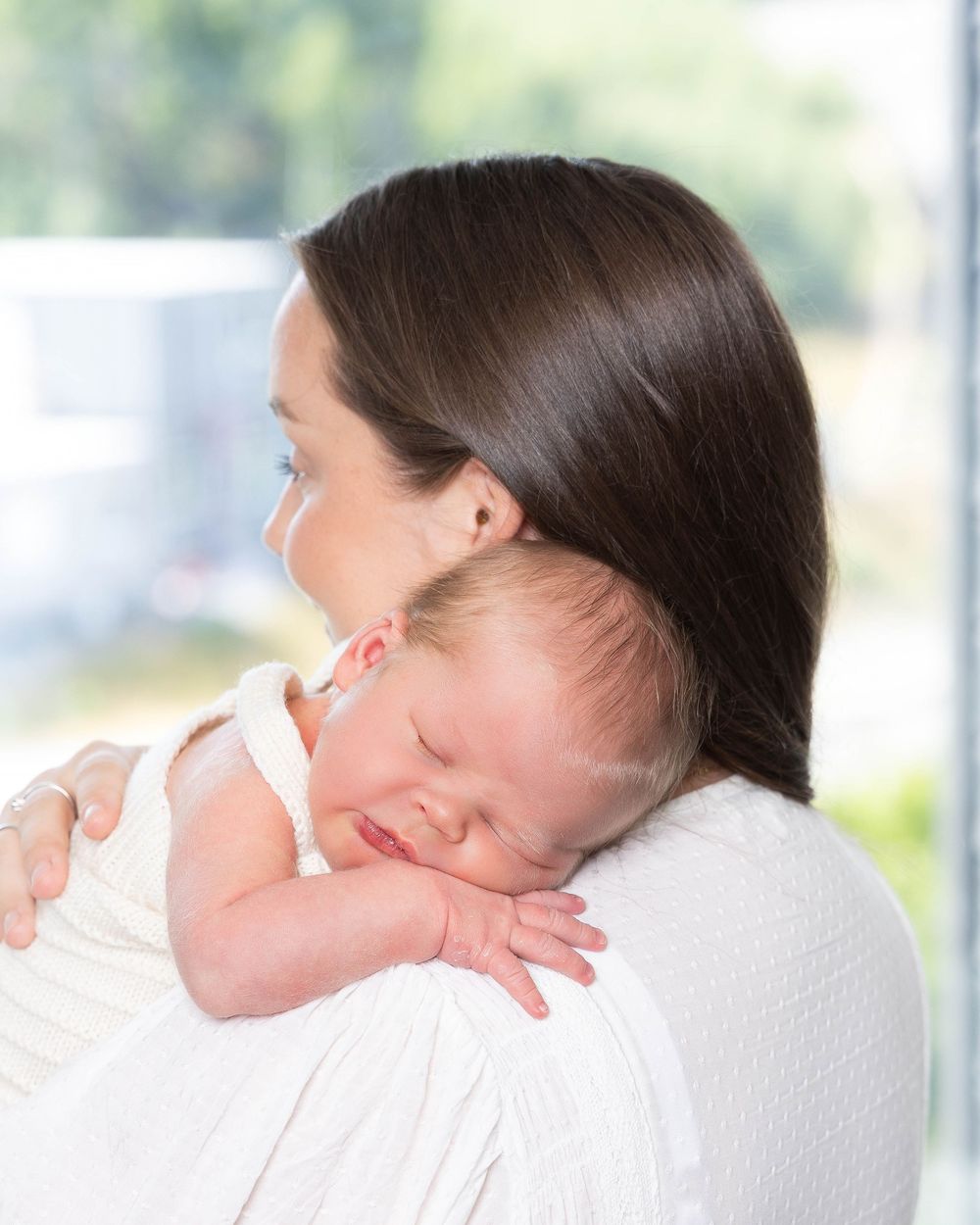 A mother in white gently holds her sleeping newborn baby on her shoulder near a bright window.