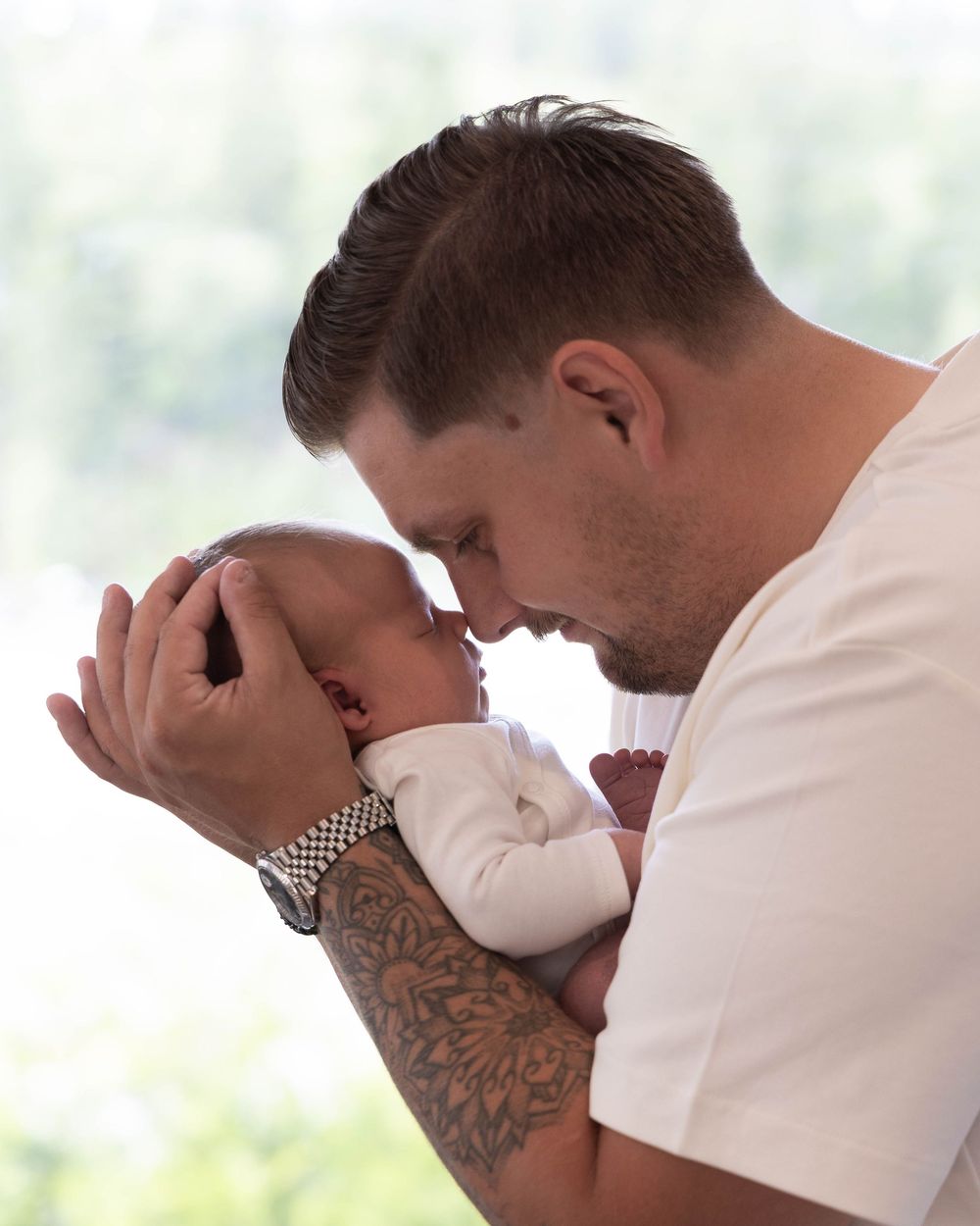 A tattooed father tenderly holds his newborn baby, touching foreheads in an intimate bonding moment.