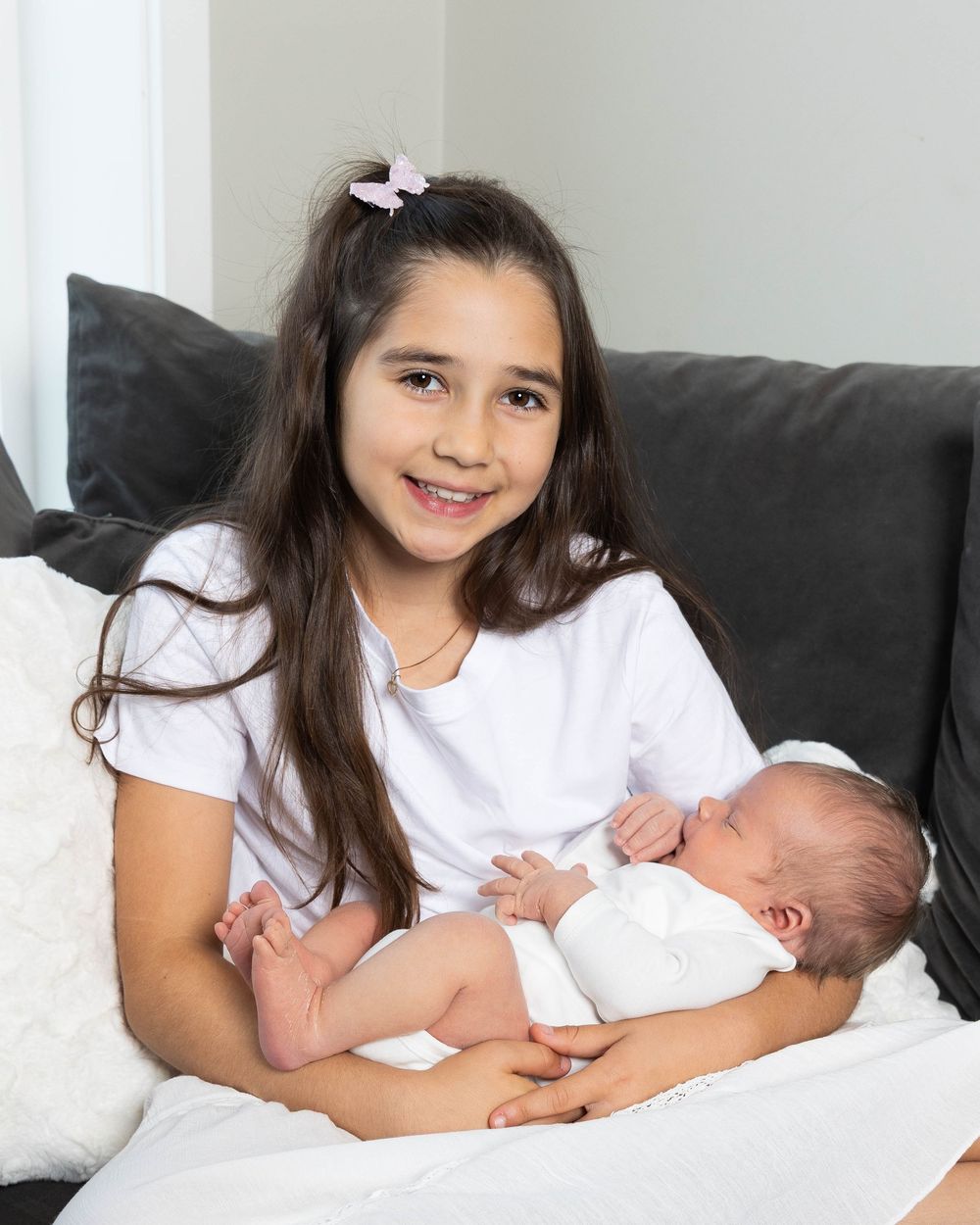 A young girl with long dark hair holds a sleeping newborn baby while sitting on a gray couch, smiling warmly.