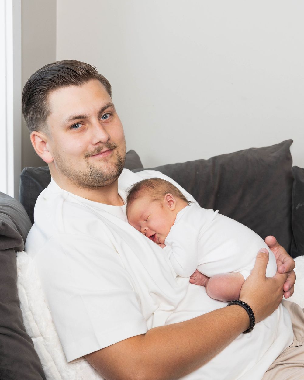 A young man smiles while cradling a sleeping newborn baby in white on a gray couch.