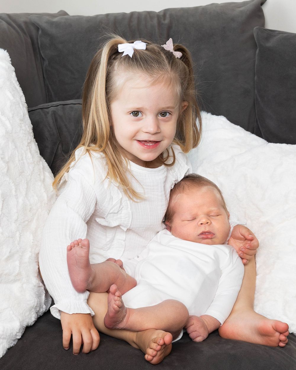 Young girl with pigtails in white dress holds sleeping newborn baby on gray couch with white blanket.