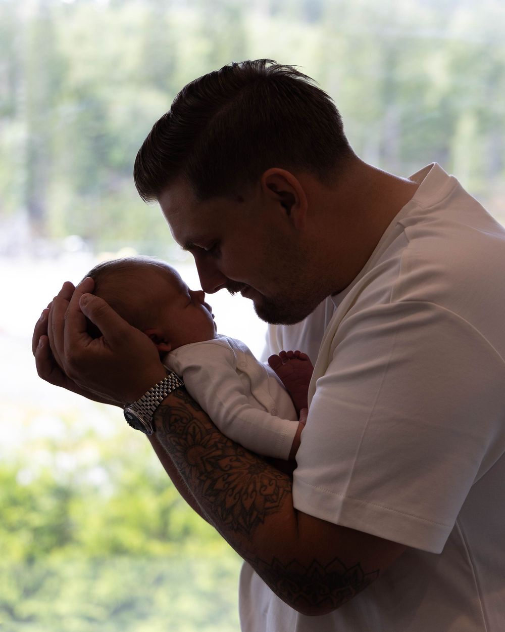 A tattooed father tenderly holds his newborn baby nose-to-nose near a window with green trees in background.