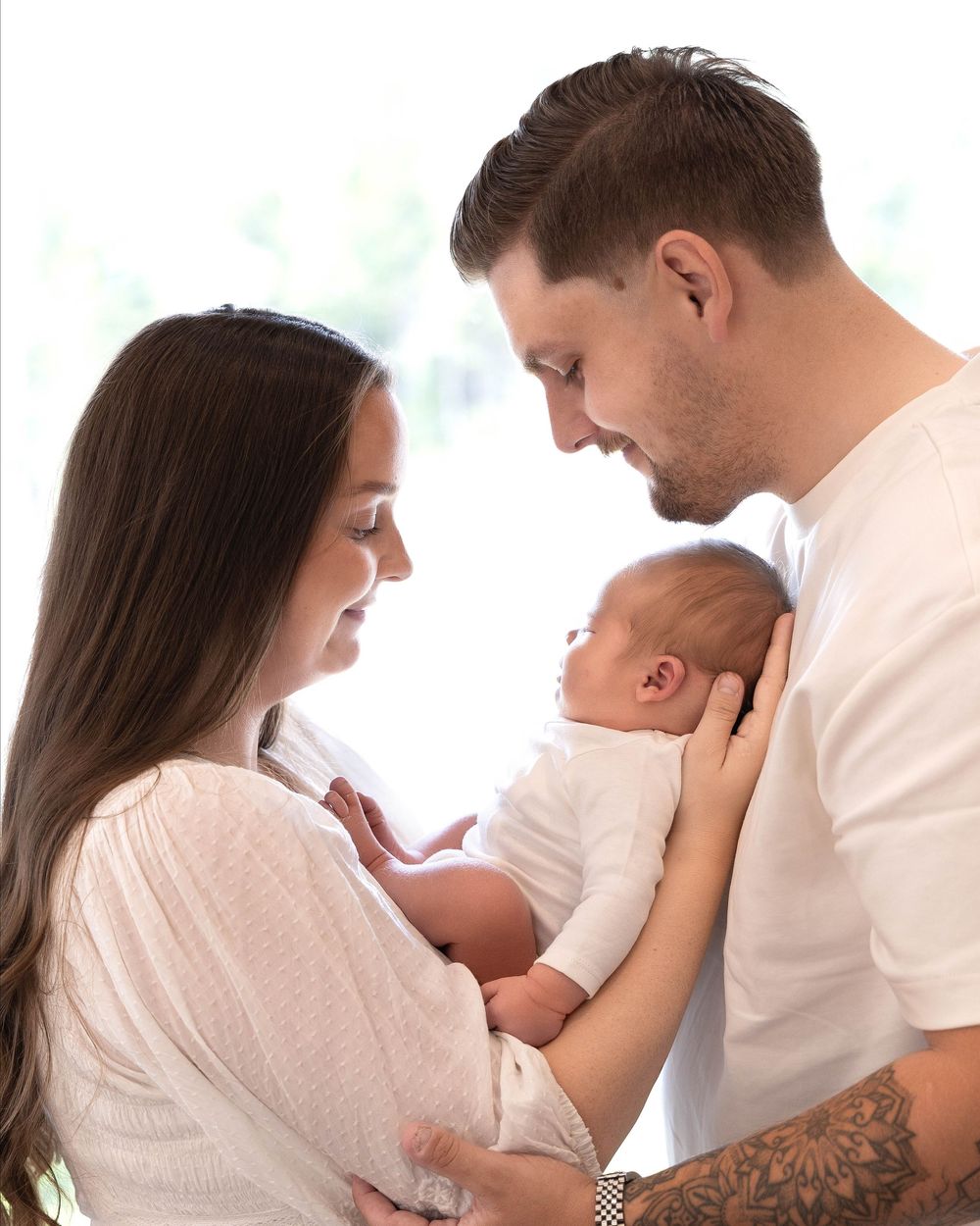 A young couple in white clothing lovingly gazes at their newborn baby, sharing a tender family moment.