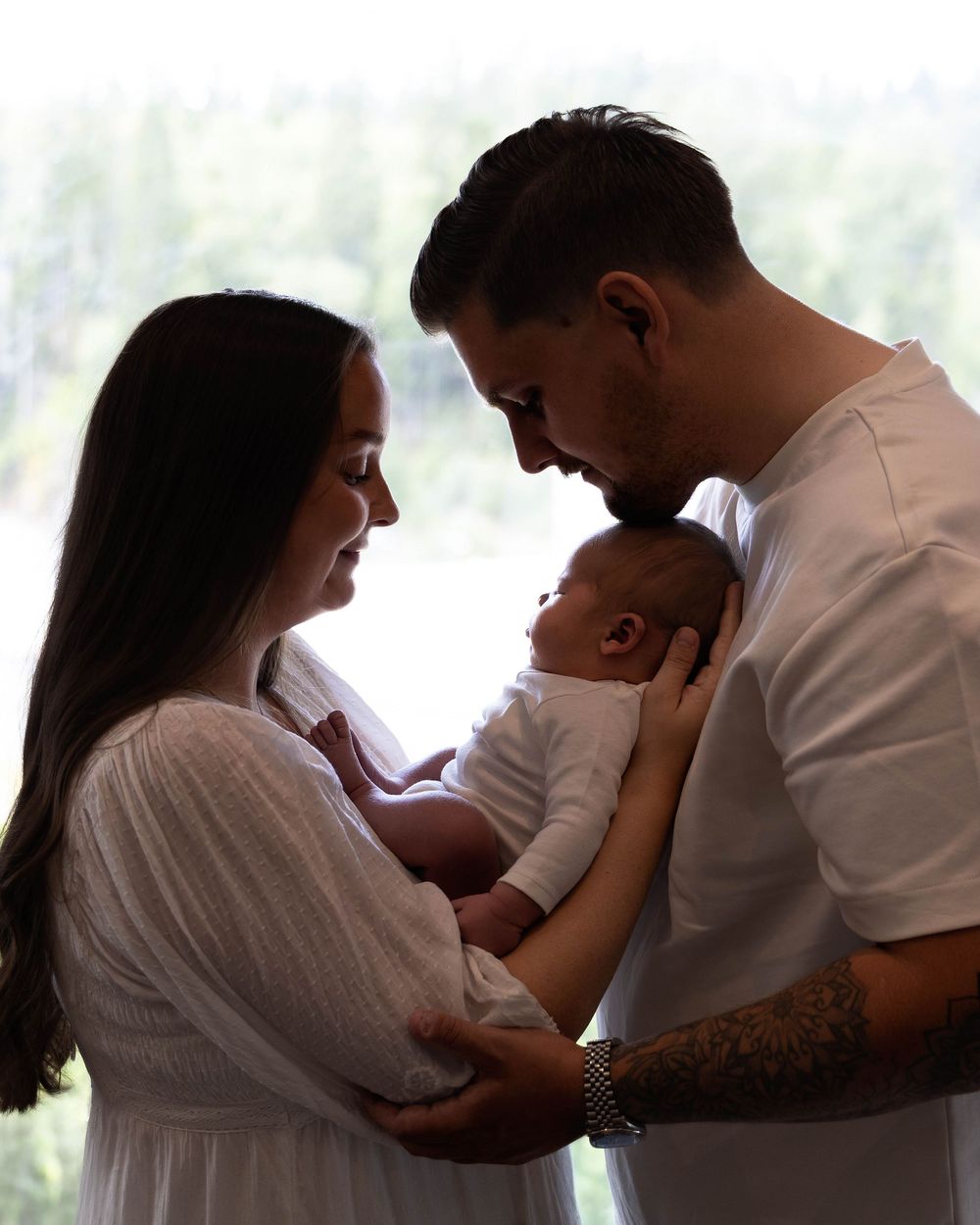 A couple tenderly holds their newborn baby together, bathed in soft natural light in an intimate family portrait.