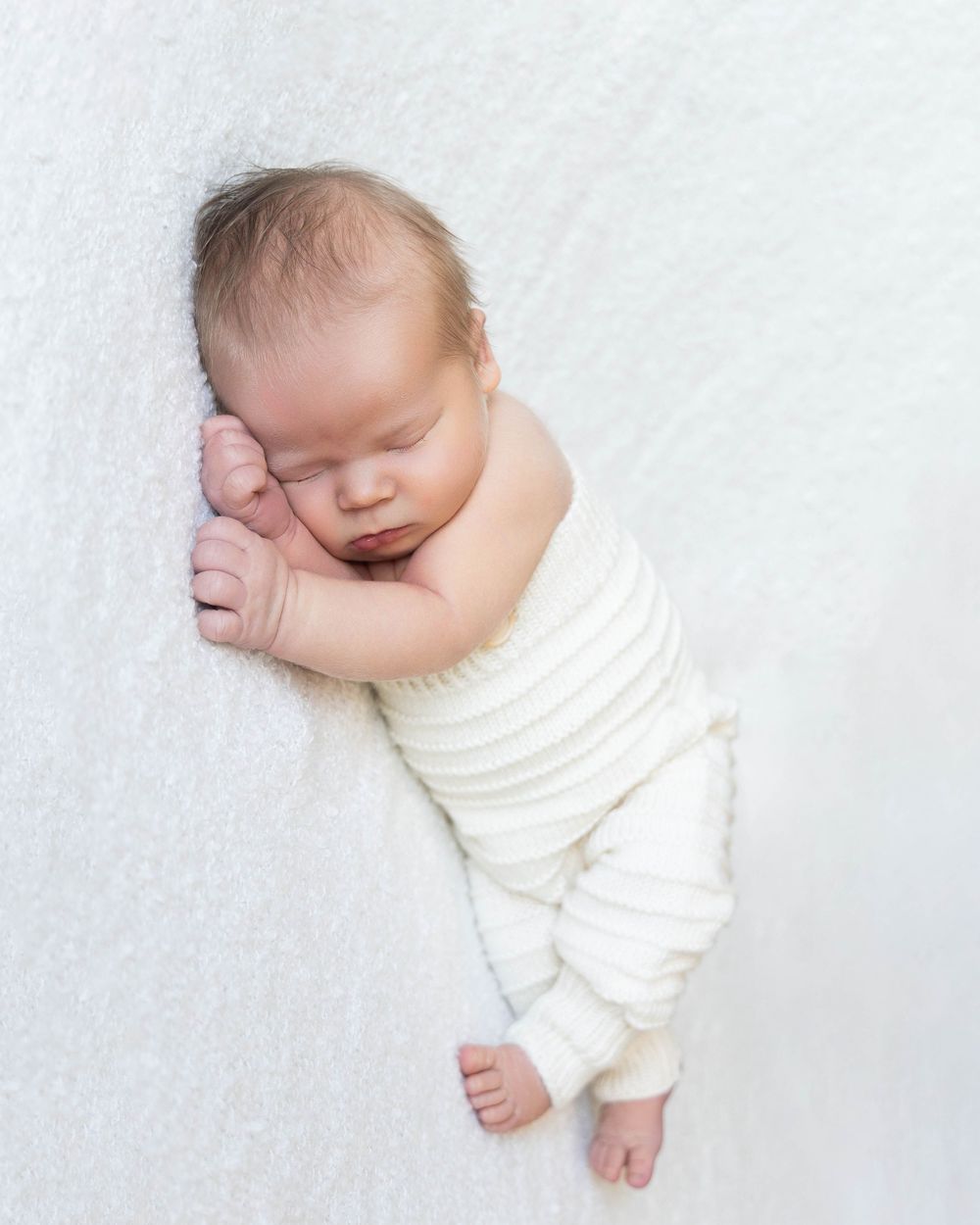 Peaceful newborn baby sleeping on white blanket wearing cream striped outfit, professional newborn photography.