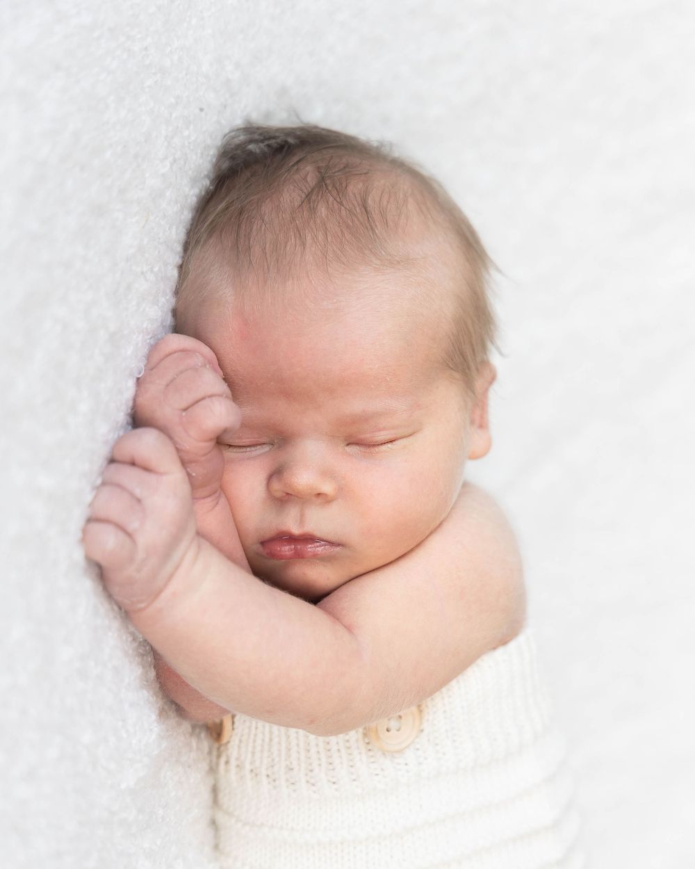 Peaceful newborn baby sleeping on white fluffy blanket, hands tucked under chin, wrapped in cream knit wrap.