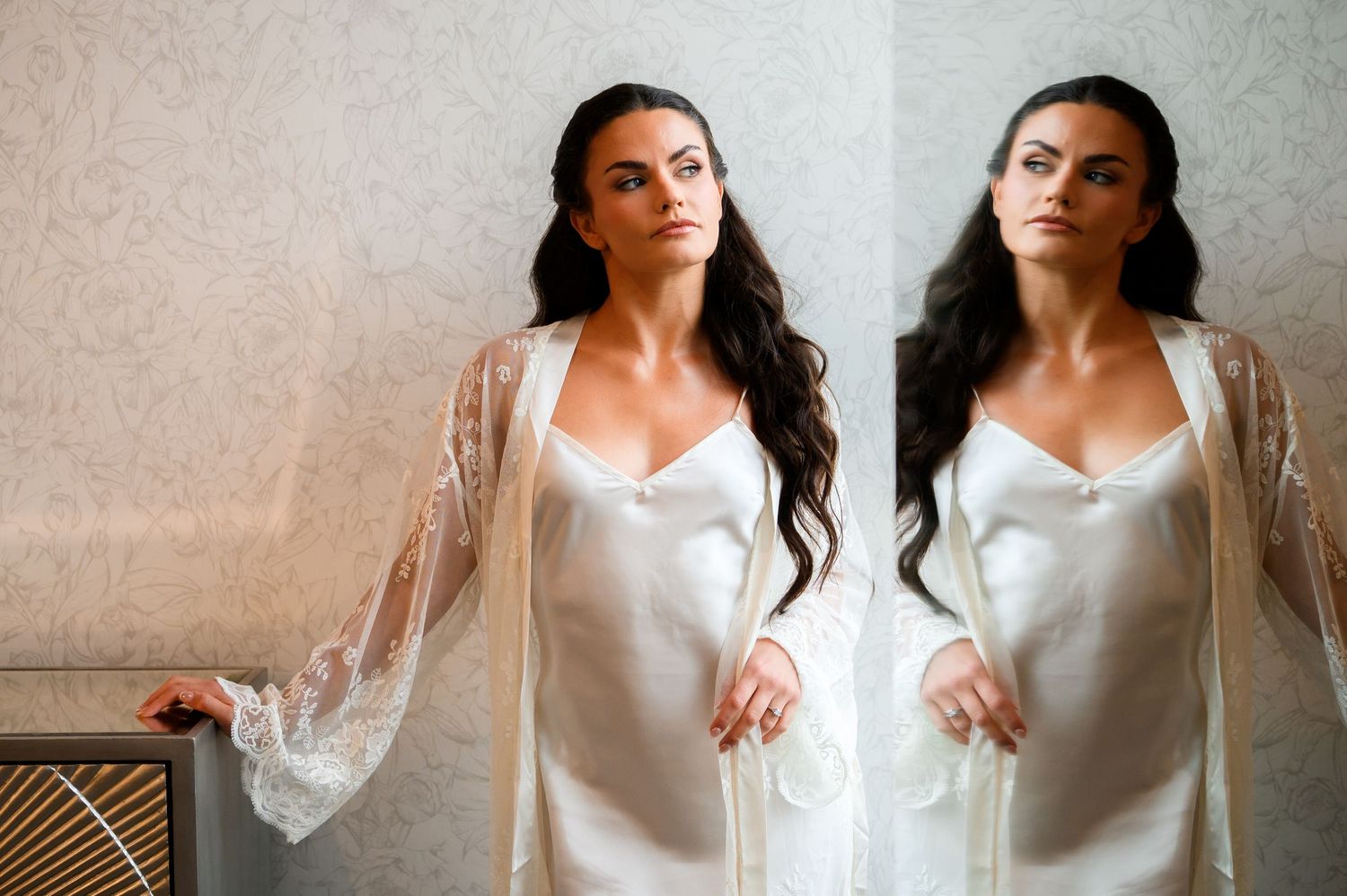 Woman in white satin slip dress and lace robe poses elegantly beside her mirror reflection in a softly lit room.