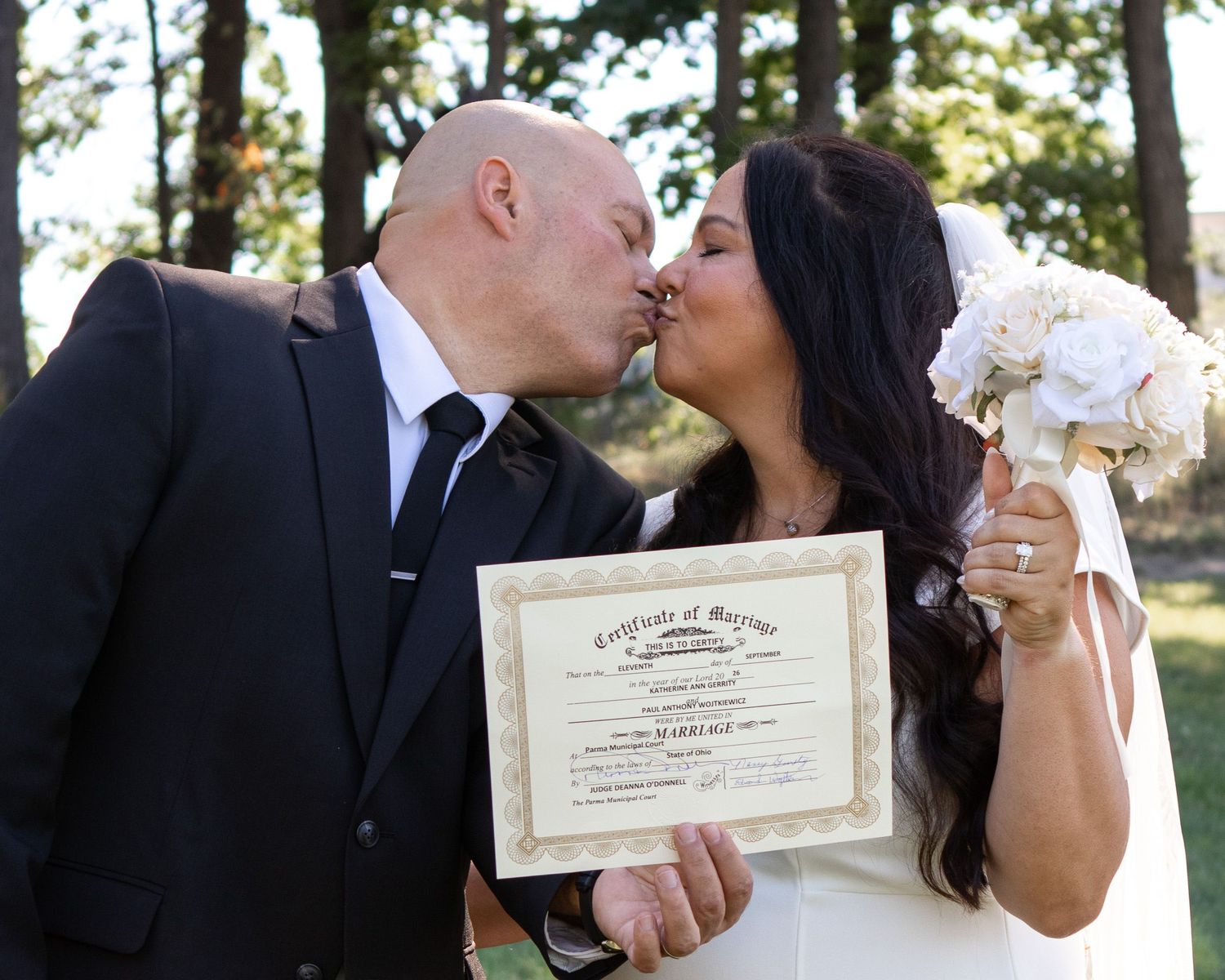 Bride in white dress and groom in black suit holding hands and smiling at each other on a green lawn.