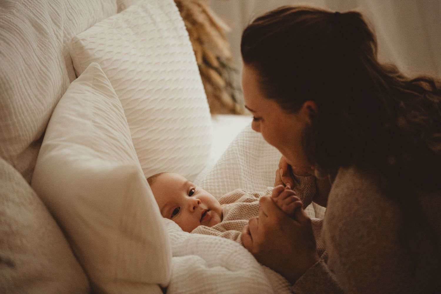 A mother tenderly gazes at her newborn baby lying on a white bed surrounded by soft pillows.