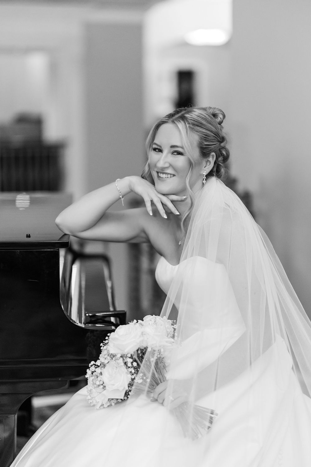 Smiling bride in strapless gown and veil leans on grand piano holding bouquet, black and white wedding portrait.