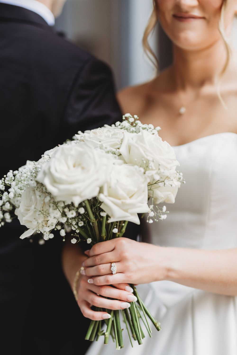 Bride in white gown holds elegant bouquet of white roses and baby's breath, diamond ring visible on her hand.