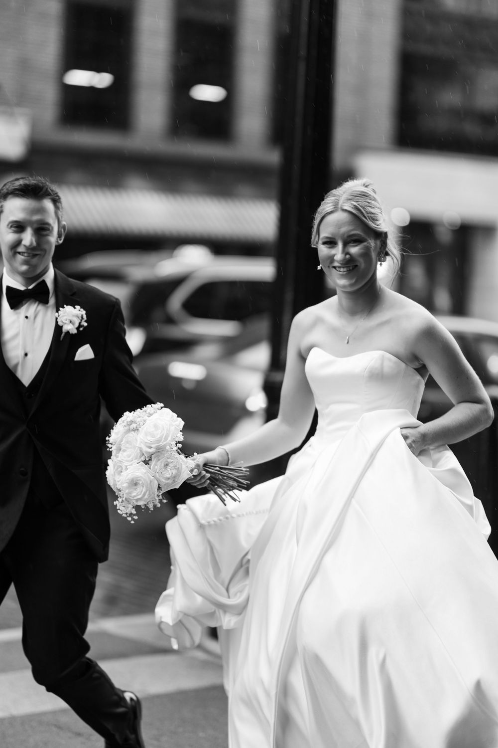 Black and white photo of smiling bride in strapless gown holding bouquet walking with groom in tuxedo on city street.