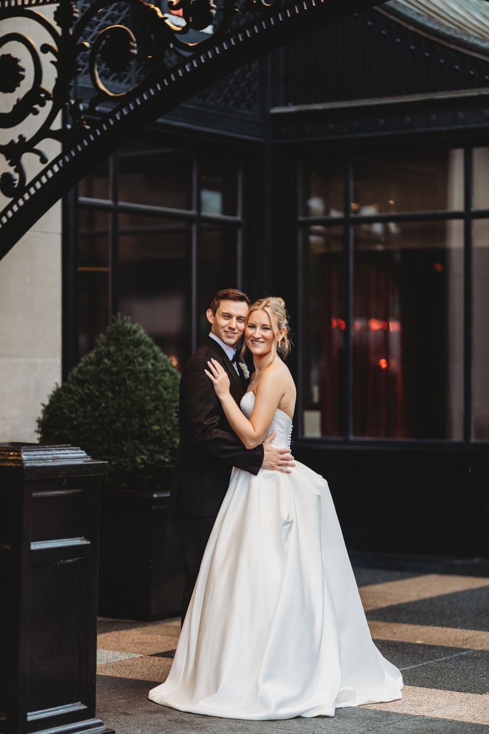 A bride in a white ballgown and groom in a black tuxedo embrace and smile outside an elegant dark building.
