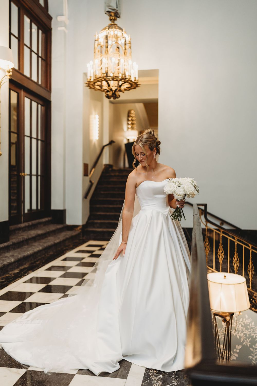 Bride in strapless white ball gown holding bouquet on grand staircase with chandelier in elegant venue.