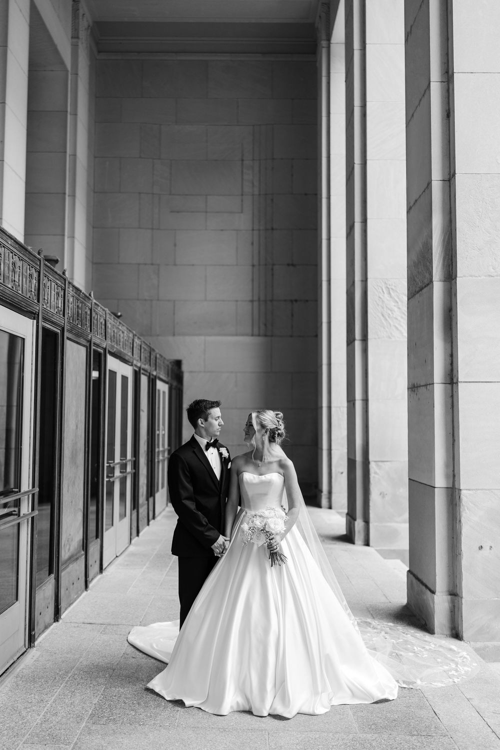 Black and white photo of bride and groom holding hands among grand columns in elegant wedding attire.