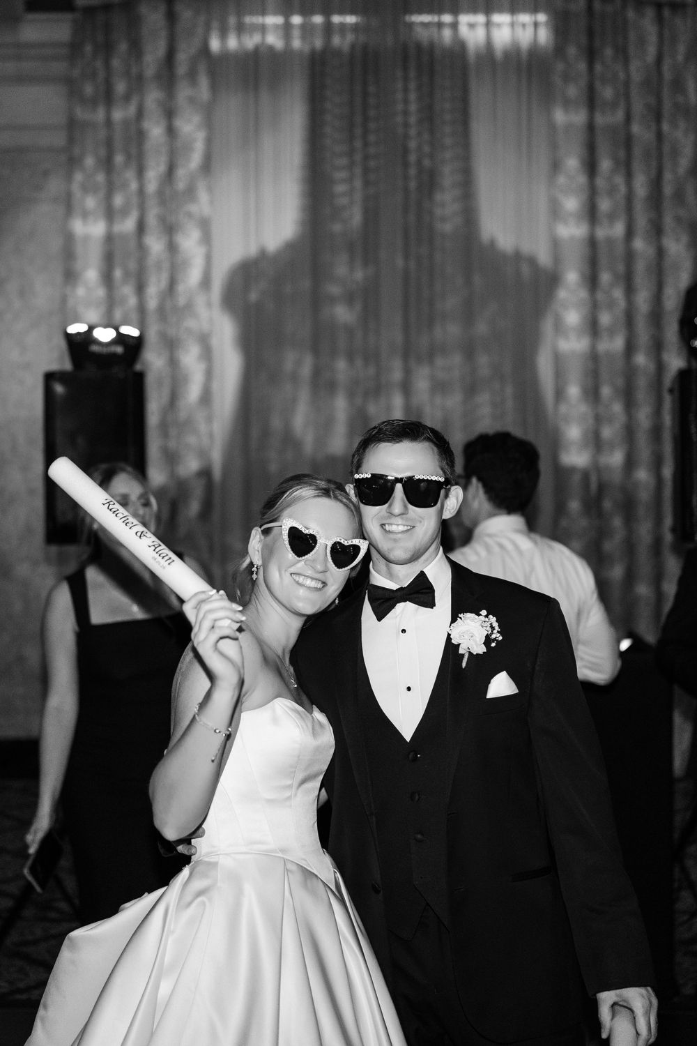 Black and white photo of a bride and groom in sunglasses posing at their wedding reception, bride holding a novelty bat.