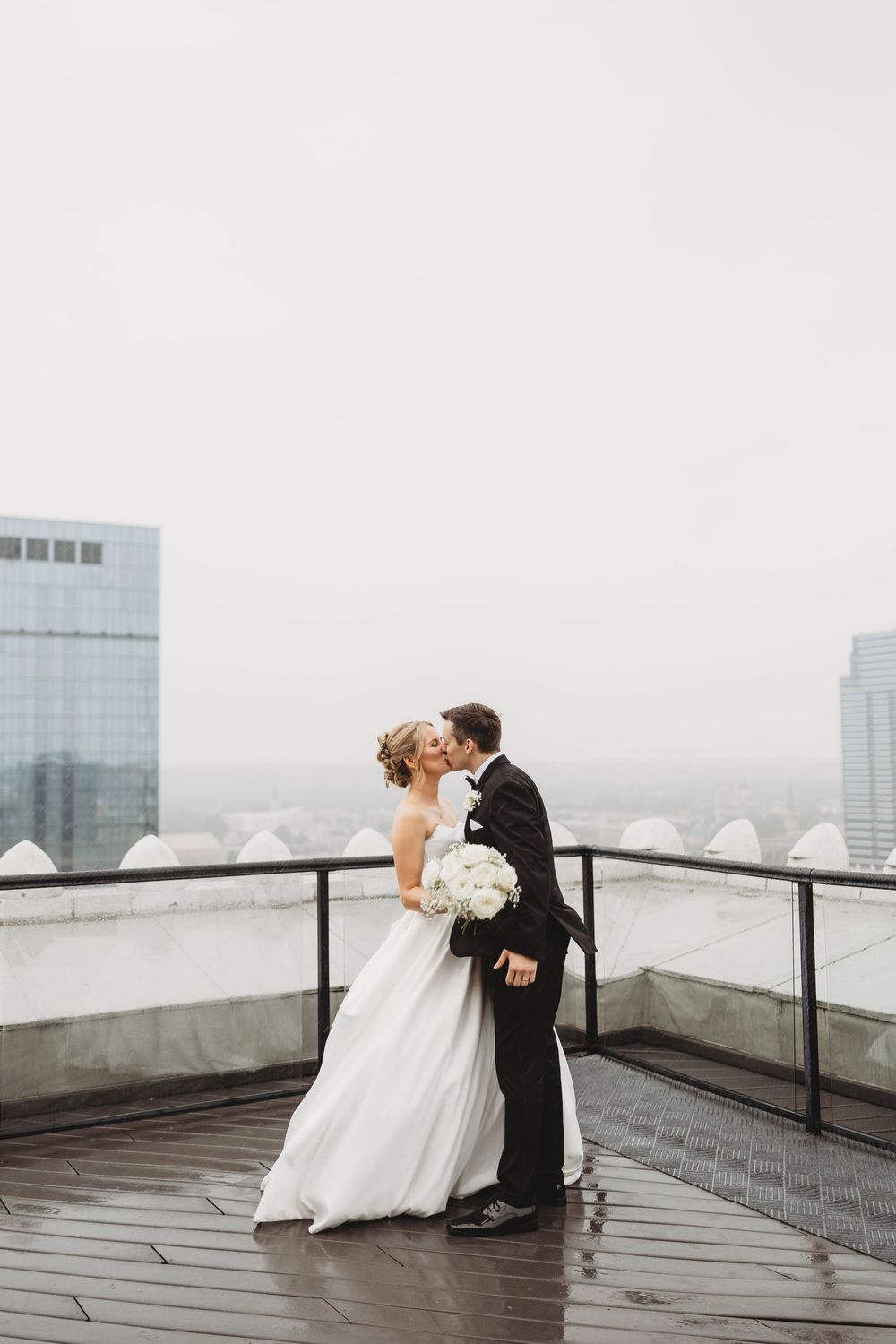 Bride and groom share a romantic kiss on a rooftop terrace, with city skyline visible in the background.