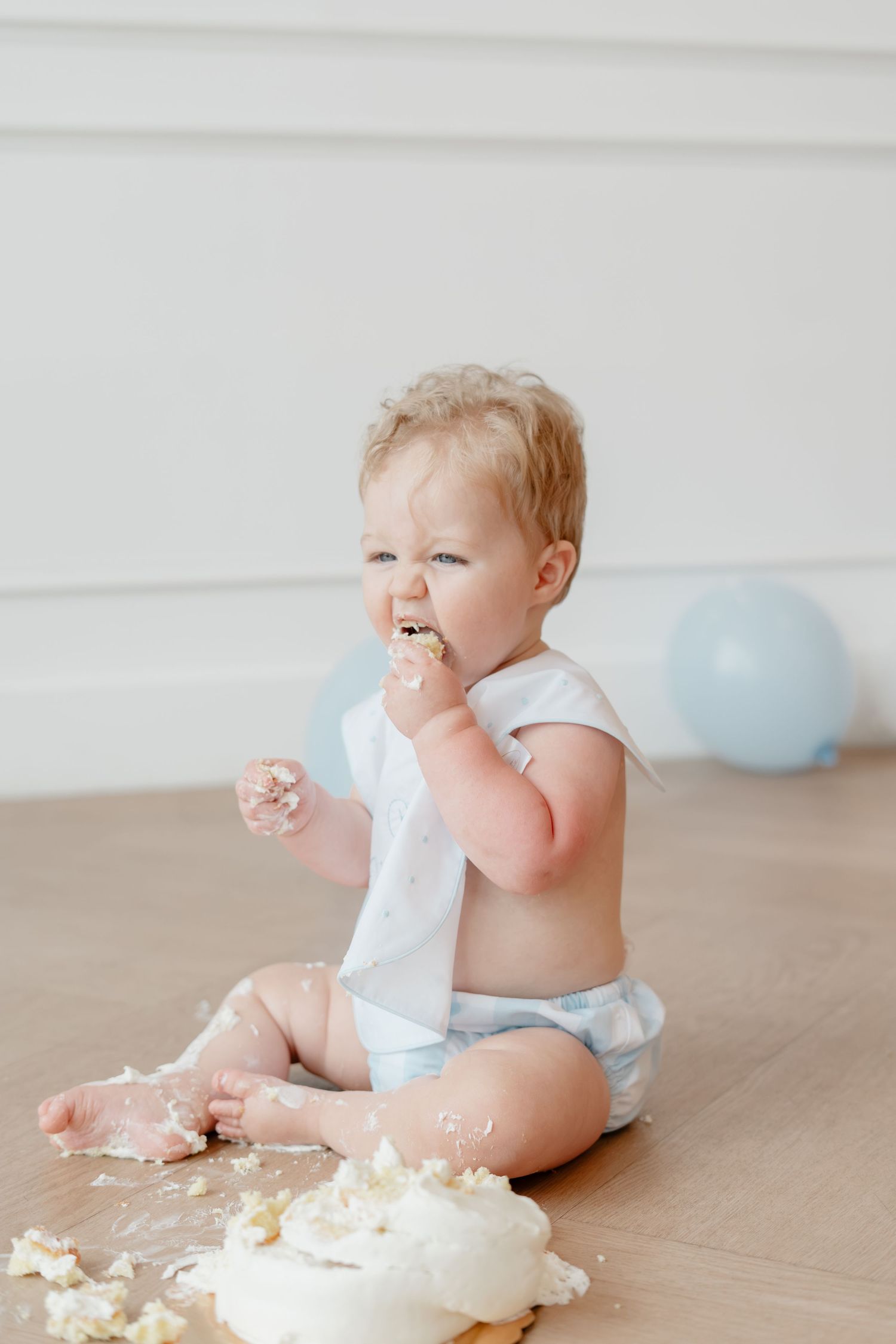 Adorable baby in diaper enjoys first birthday cake smash, sitting on floor with frosting on face and hands.