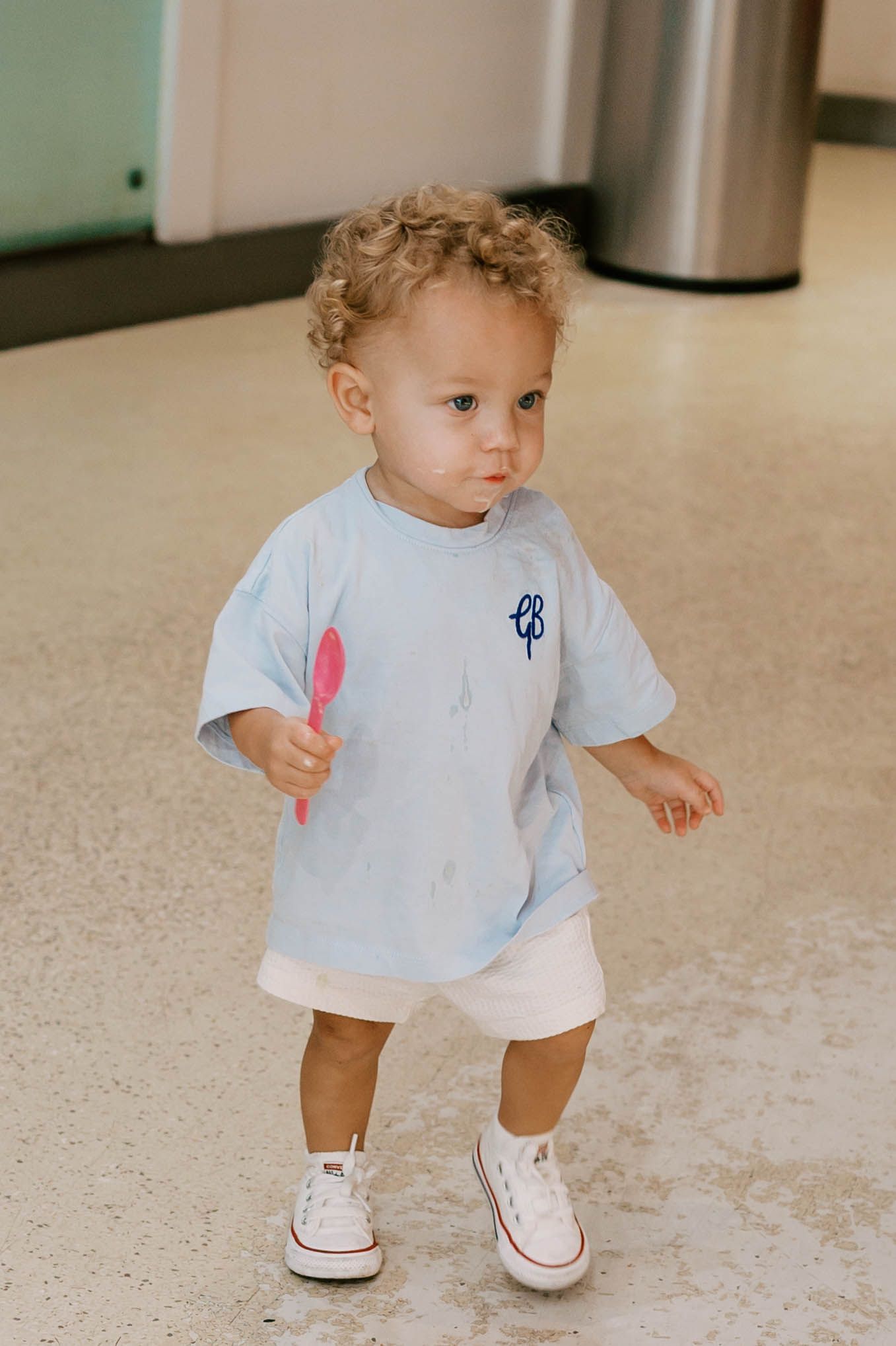 Cute toddler in an oversized light blue shirt and white shorts walks while holding a pink spoon.