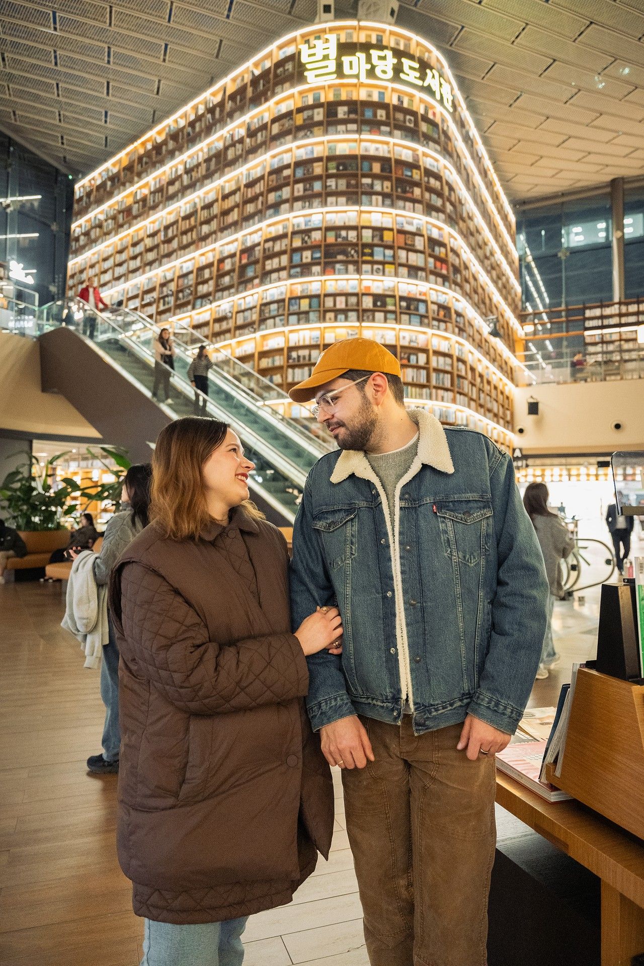 Couple holding hands at Starfield Library in Seoul, South Korea, with illuminated bookshelves behind them.