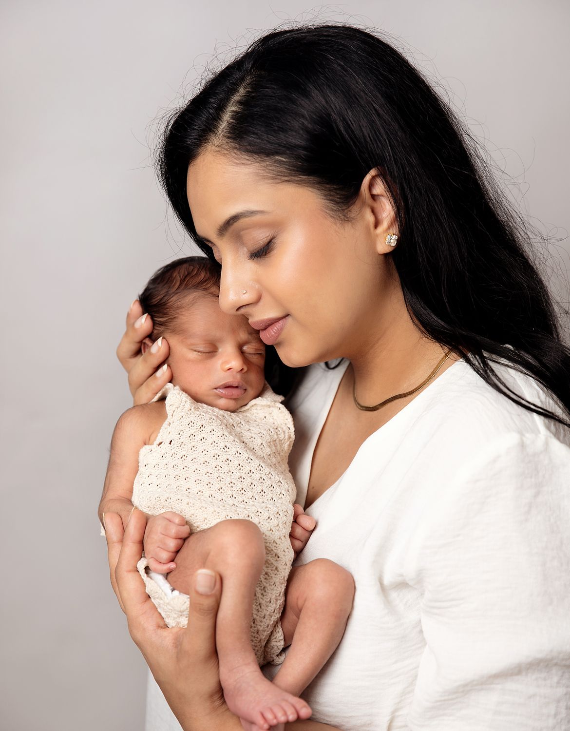 A mother holds her sleeping newborn baby wrapped in a cream knit swaddle against a white background. During a newborn session in Orpington